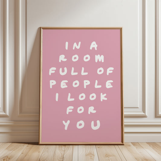 An unframed typographic wall art print. The print has white hand-drawn text reading “IN A ROOM FULL OF PEOPLE I LOOK FOR YOU” on a  pink background.